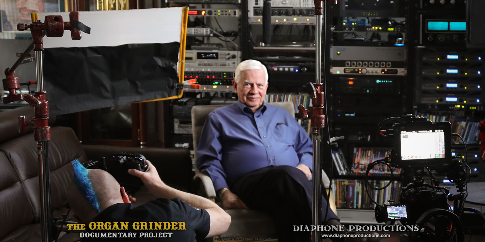 Behind-the-scenes production photo. Dennis Hedberg is seated in front of his wall-sized audio system, surrounded by camera and lighting equipment.