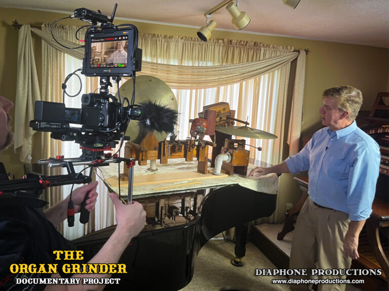 Behind-the-scenes photo of Don Feely being interviewed on-camera. Don is standing beside his grand piano. On top of the piano, an array of drums, percussions, and traps from the original Organ Grinder are on display.