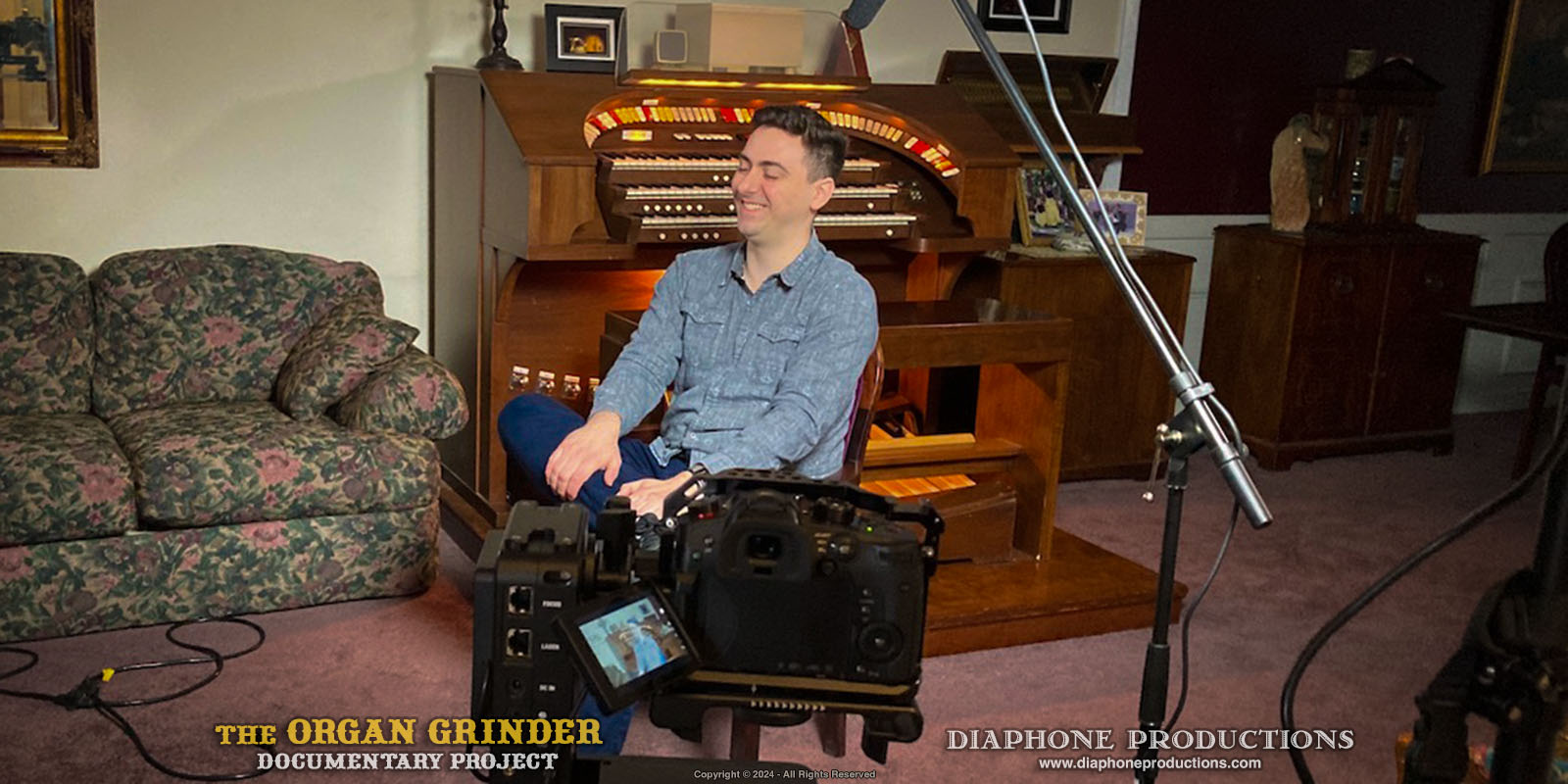 Nathan Avakian in front of the camera about to be interviewed. He is sitting in a small chair in his parent's living room, in front of his childhood practice organ console.