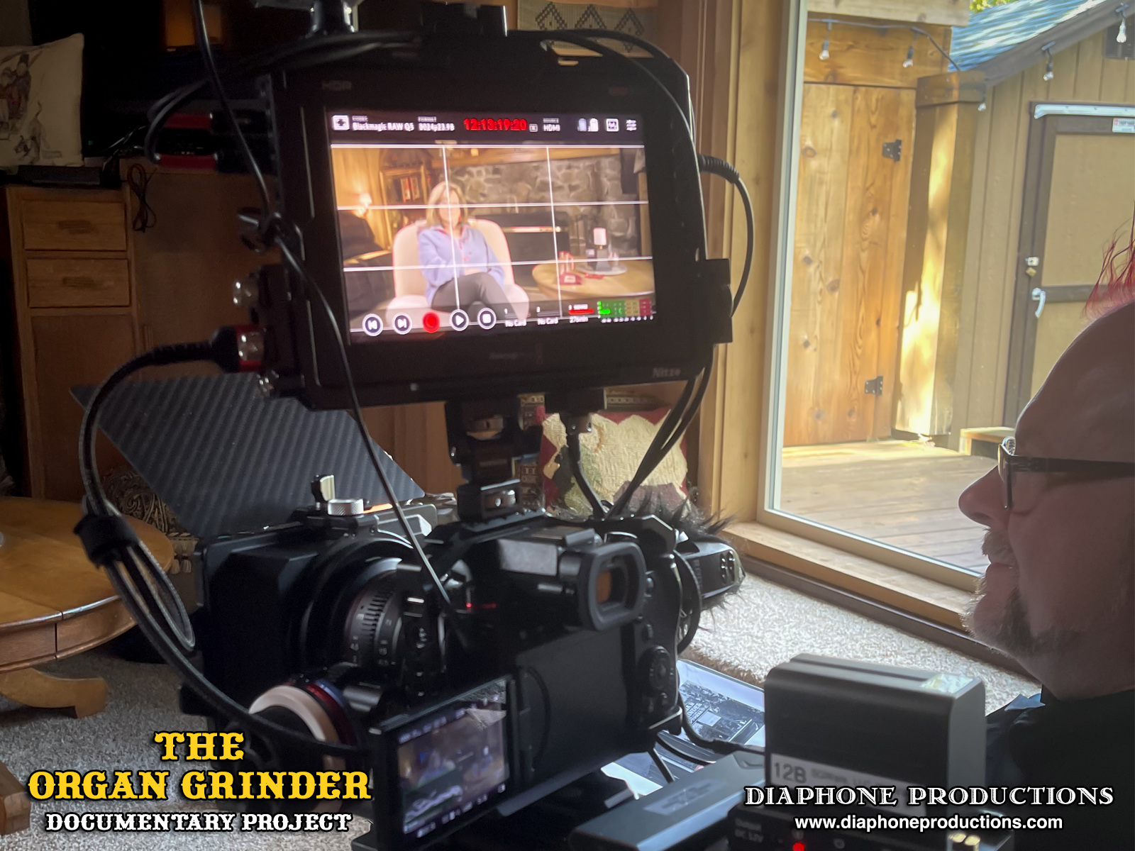 A behind-the-scenes photo of a cinema camera rig in Vicki Buck's rustic cabin living room. The camera monitor shows Vicki sitting in a chair.
